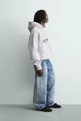 Sudadera con capucha y bolsillo frontal tipo canguro, confeccionada en tejido de algodón con interior perchado. Presenta diseño de corte boxy fit, mangas largas con puños y ruedo en rib, y texto bordado y estampado en el frente.