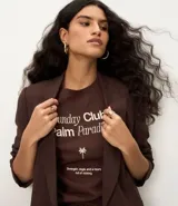 Remera de algodón color marrón oscuro, con cuello redondo y manga corta. Presenta un estampado frontal en blanco con el texto "Sunday Club Palm Paradise", una palmera y una frase motivacional.