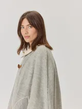 Capa de tejido de punto en color gris melange, con diseño abierto y cierre frontal mediante un botón tipo duffle de madera con detalle en cuero. Presenta bolsillos laterales amplios y bordes con costuras a la vista.