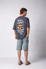 Remera de corte oversize color gris oscuro, confeccionada en algodón premium de alto gramaje. Presenta un diseño minimalista con un pequeño estampado frontal que incluye un oso y el texto 'Richer & Louder VCP'.