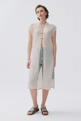 Kimono tejido de punto en color crudo con sutil brillo de lurex. Presenta un diseño sin mangas, corte largo y cierre frontal mediante dos lazos finos. El tejido tiene una textura acanalada vertical.