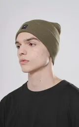 Gorro de tejido de punto color verde militar, de estilo beanie, con borde doblado y un pequeño parche negro con un logo de tijeras cruzadas.
