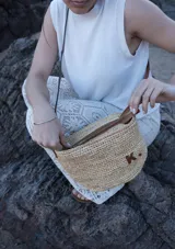 Bandolera de rafia natural de forma redondeada, con correa larga fina de cuero marrón, cierre superior y detalle de aplique en cuero con la letra K.