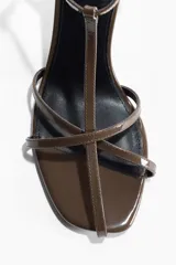 Sandalias de taco alto con acabado charolado, color marrón, diseño de tiras finas con tira en T y pulsera ajustable con hebilla metálica en el tobillo.