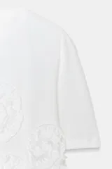 Camiseta de algodón de corte regular, con cuello redondo y manga corta. Presenta un detalle decorativo de flores con volumen confeccionadas en el mismo tejido sobre el frente.