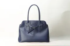 Cartera shopper negra de cuero sintético con asas largas para el hombro, detalles de correas frontales con hebilla y marca She Mona grabada en el frente.