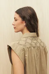 Camisa color beige, de corte recto, con cuello clásico, mangas cortas con volados en los hombros y cierre frontal con botones.