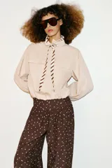 Pantalón de tiro alto con pernera ancha y diseño fluido. Presenta un estampado de lunares blancos sobre fondo marrón y detalle de frunces en la cintura.