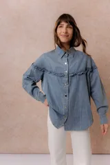 Camisa de denim liviano con calce relajado, cuello camisero clásico y cierre frontal con broches metálicos. Presenta un canesú con volado en el delantero y la espalda, mangas largas con puños abotonados y terminación inferior redondeada.