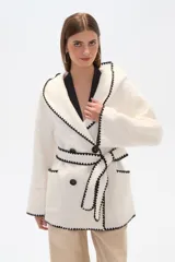 Chaqueta blanca de corte holgado con capucha, cierre frontal cruzado con botones y cinturón de tela ajustable. Presenta detalles de costuras decorativas en color negro en los bordes, solapas, bolsillos y cinturón.