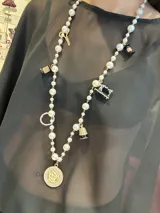 Collar largo de perlas blancas con dijes colgantes de metal dorado y esmalte, incluyendo motivos de costura como tijeras, carretes de hilo y una máquina de coser, finalizando en un dije circular central con diseño de flor.