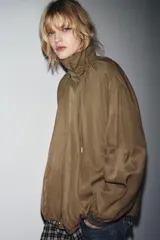 Chaqueta tipo bomber fluida color marrón, con cuello alto ajustable con cordones y stoppers, manga larga con elástico en los puños, bolsillos delanteros y bajo con pliegues. Cierre frontal con cremallera y botones a presión ocultos por solapa.