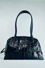 Bolso de hombro de piel con formato bowling, acabado texturizado tipo cocodrilo y color bordó. Cuenta con doble asa de hombro, cierre principal de cremallera con doble tirador y bolsillo interior con cremallera.