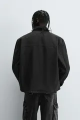 Cazadora de algodón color negro lavado, de corte relajado (relaxed fit), con cuello solapa a contraste, cierre frontal de cremallera, bolsillos canguro en la cadera y puños con botón a presión.