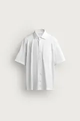 Camisa blanca de manga corta con corte regular fit, confeccionada en tejido fluido de algodón y lino. Presenta cuello solapa, cierre frontal de botones y un bolsillo de parche en el pecho.