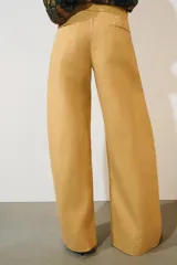 Jeans de tiro bajo y corte wide leg, color amarillo mostaza, con pliegues frontales, bolsillos ocultos y cierre frontal con cremallera, botón y ganchos metálicos.