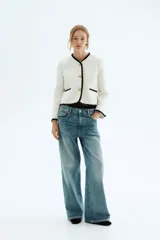 Chaqueta corta de tejido bouclé blanco, con ribetes negros en cuello, frente, bolsillos y puños. Presenta cuello en V, cierre frontal con botones dorados con diseño de nudo y bolsillos insertados.