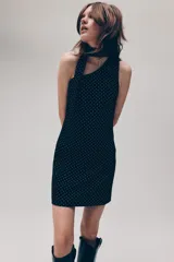 Vestido corto negro con estampado de lunares blancos, escote asimétrico de un solo hombro y tira ancha decorativa que cae desde el hombro.