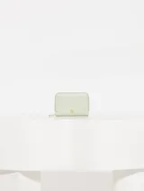 Billetera pequeña con cierre de cremallera, fabricada en cuero vacuno graneado de color celeste pálido. Presenta un logo de marca gofrado en el frente y herrajes dorados.