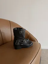Bota de caña media en eco cuero negro, con diseño estilo biker que incluye correas con hebillas metálicas y herrajes plateados en el tobillo. Cuenta con suela plana y punta redondeada.