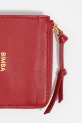 Monedero o tarjetero pequeño de cuero rojo, con cierre de cremallera superior y un tirador de tela a juego. Presenta un bolsillo exterior frontal y el logo de la marca grabado en dorado.