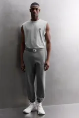Pantalón jogger gris de corte regular fit, confeccionado en tejido de algodón con interior afelpado. Cuenta con cintura elástica ajustable con cordón, bolsillos laterales de vivo y traseros de plastrón. Presenta un texto estampado en color bordó en la pernera y bajo acabado en puño con elástico. Incluye un parche con logo combinado a contraste en la cintura.