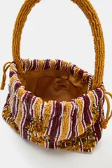 Bolso de hombro de tamaño mini, confeccionado con cuentas y abalorios que forman un diseño a rayas verticales y detalles de flores bordadas en tonos mostaza, bordó y blanco. Cuenta con asa corta de mano y cierre fruncido con cordones.
