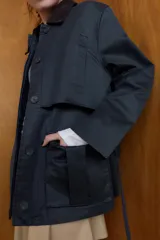 Chaqueta encerada de corte recto con cuello de solapa en pana marrón. Presenta manga larga, bolsillos delanteros de vivo y de plastrón con solapa, y un cinturón de lazada en el mismo tejido. Incluye cierre frontal con botones ocultos por tapeta y abertura en la espalda.