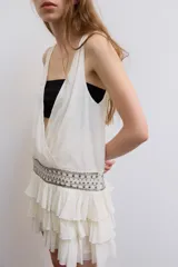 Vestido corto de gasa color blanco, con escote en V cruzado, sin mangas y diseño con volantes en el bajo. Presenta un detalle de tachas plateadas en la cintura.