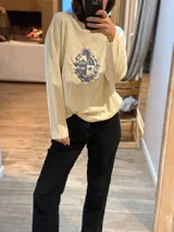 Sweater de punto color crema, de corte holgado y manga larga, con cuello redondo. Presenta un escudo bordado en azul en el centro del frente.