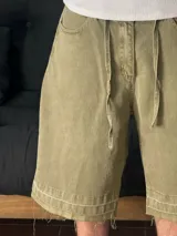 Bermuda de corte baggy color verde con efecto gastado. Presenta costura vertical trasera en cada pierna y detalle artesanal visible en el bajo. Incluye un cordón extra confeccionado en la misma tela.