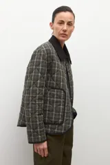 Campera capitoneada de corte recto con diseño a cuadros en tonos verde oliva y gris. Presenta cuello camisero de pana en color negro, cierre frontal con botones a presión y bolsillos laterales amplios con ribete negro.