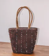 Cartera tipo tote de gamuza color marrón, con correas trenzadas y detalle de piedras naturales incrustadas en líneas verticales. Cuenta con cierre metálico y forro interior de tela.