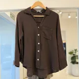 Camisa marrón oscuro de manga larga, con cuello camisero, cierre frontal de botones claros, un bolsillo aplicado en el pecho y dobladillo asimétrico más largo en la parte trasera.
