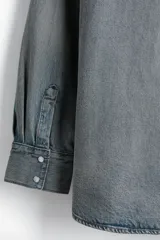 Camisa vaquera de algodón rígido, color celeste lavado, con cuello, botones a presión frontales y en los puños, canesú delantero y trasero, y dos bolsillos superiores con solapa y botón a presión. Presenta un bajo redondeado y un corte oversized.