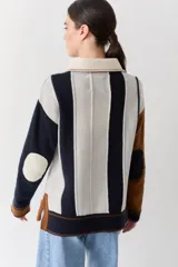 Buzo de punto con cuello polo y cierre frontal de tres botones. Presenta un diseño a rayas verticales en tonos beige y marrón, con mangas en bloques de color rojo y detalles en rosa en los puños y el ruedo.