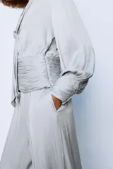 Camisa de escote en pico con lazada frontal en el mismo tejido, manga larga con puños y bajo drapeado. Presenta un acabado satinado y cierre lateral con cremallera oculta.