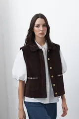 Chaleco de pana color marrón oscuro, con cuello camisero, cierre frontal de botones metálicos dorados y dos bolsillos frontales con detalle de bordado geométrico.