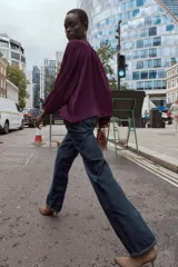 Blusa de tejido semitransparente color violeta, con cuello redondo y detalle de fornitura dorada en el escote. Presenta un diseño de escote en pico, mangas largas abullonadas con puños elásticos y corte holgado.