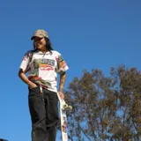 Llavero con tira de tela a rayas horizontales en tonos celeste, blanco, naranja, rosa y verde. Tiene un mosquetón metálico color bronce y una pieza de cuero marrón con el logo de la marca.