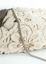 Cartera clutch color beige de crochet con diseño floral y aplique de caracoles en el centro de una de las flores.