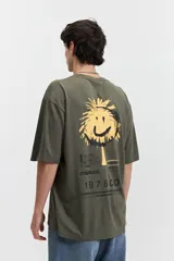 Remera oversize verde militar de manga corta y escote redondo, con estampado trasero de una cara sonriente amarilla con detalles en negro y texto "mistral 1976co".