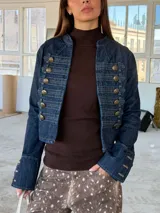 Chaqueta de denim estilo militar con diseño napoleónico, caracterizada por su doble fila de botones metálicos decorativos en el frente y mangas acampanadas con detalles de botones en los puños. Presenta un corte entallado y cuello redondo.