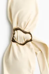 Top de bikini strapless de color beige, con copas acolchadas, varillas laterales y ribete de silicona en el escote. Cuenta con un detalle metálico dorado en el centro con tiras colgantes y ajuste en la espalda.