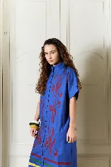 Vestido largo verde militar de popelina de algodón, con diseño oversized, cuello mao, mangas cortas, bolsillos laterales y cierre frontal con botones. Presenta un estampado de flores grandes en la parte superior y franjas horizontales de colores en el ruedo.