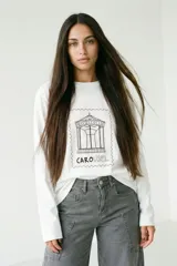 Remera de manga larga color blanco, con cuello redondo y estampa frontal que representa un carrusel dentro de un marco de sello postal.