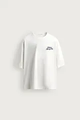 Camiseta blanca de corte relajado, con cuello redondo, manga corta y texto estampado en contraste en el pecho.