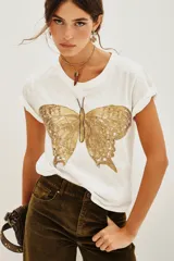 Remera de algodón color blanco con cuello redondo y mangas cortas con dobladillo. Presenta un bordado detallado de una mariposa en tonos dorados en la parte frontal.