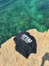 Remera marrón de corte holgado con estampado en la espalda que incluye la palabra "ITTAI" en blanco con líneas rojas y azules, la frase "AN IBIZA PRODUCE" y un texto más pequeño que dice "Ittai Stars Department Gallery - Ibiza Edition. If you're in Ibiza, 112 is not an option.".