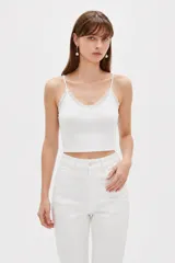 Musculosa de punto blanco, corte crop, con tirantes finos, escote en V adornado con detalles de encaje y un pequeño lazo en el centro.